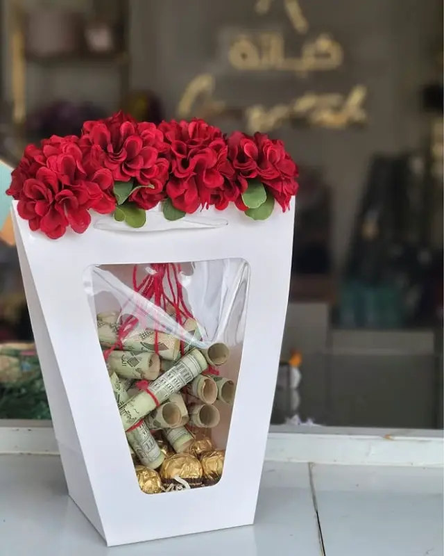 فازة عيدية من كياتة في صنعاء — فازة بيضاء يعلوها ورد أحمر وفيها فلوس ملفوفة وشوكولاتة